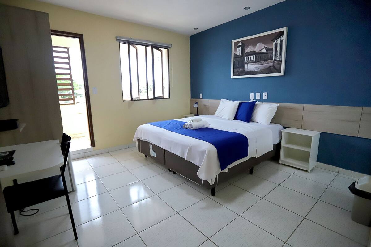 A hotel room with a bed, a desk, a chair, a window, and a wall-mounted picture.