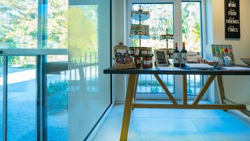 A room with a table displaying various food items and wine bottles, large windows with a view of greenery, and a wall with a sign.