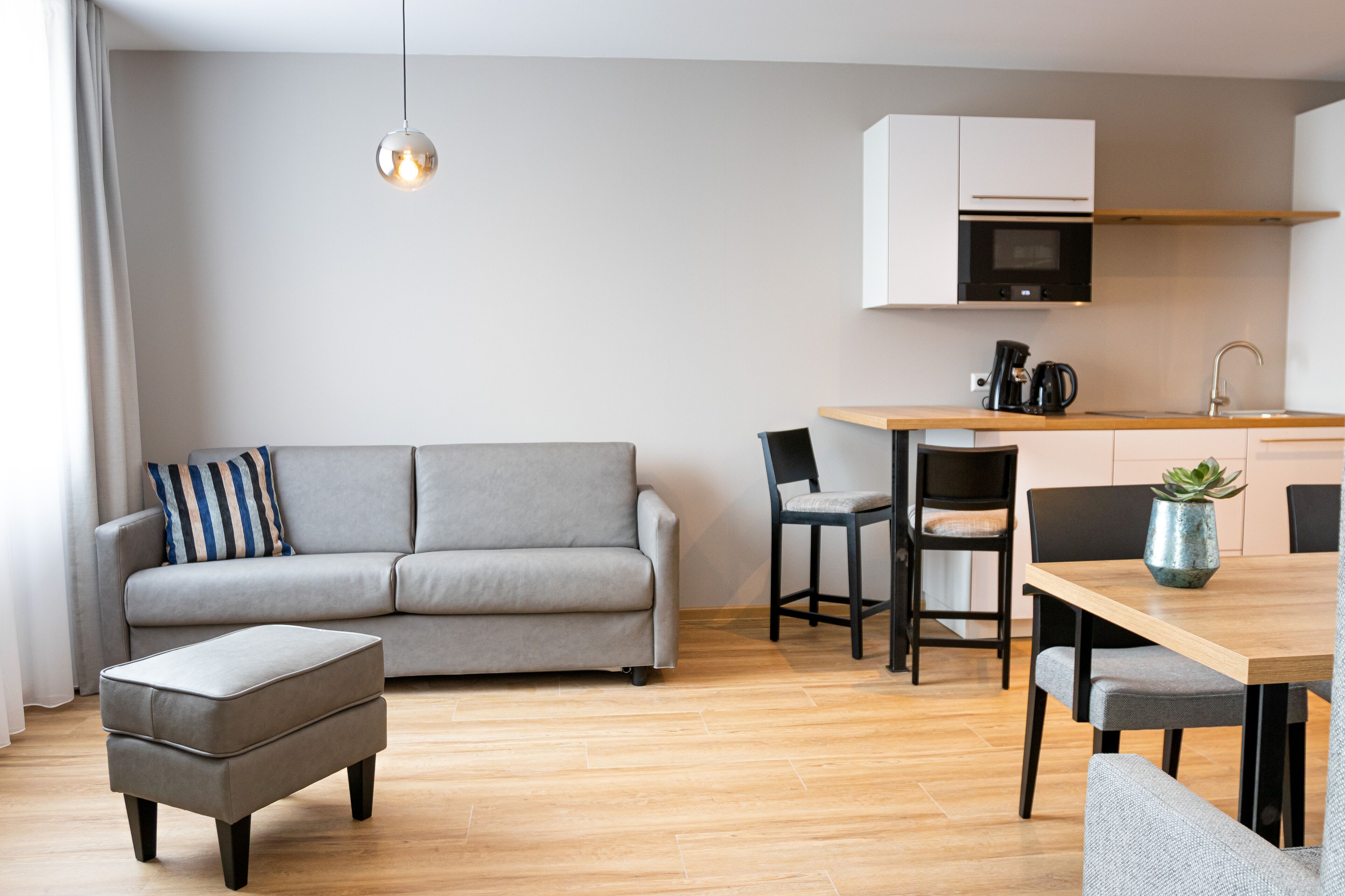 A modern living room with a grey sofa, a wooden dining table, and a kitchen area with a microwave and coffee maker.