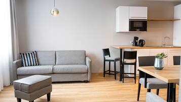 A modern living room with a grey sofa, a wooden dining table, and a kitchen area with a microwave and coffee maker.