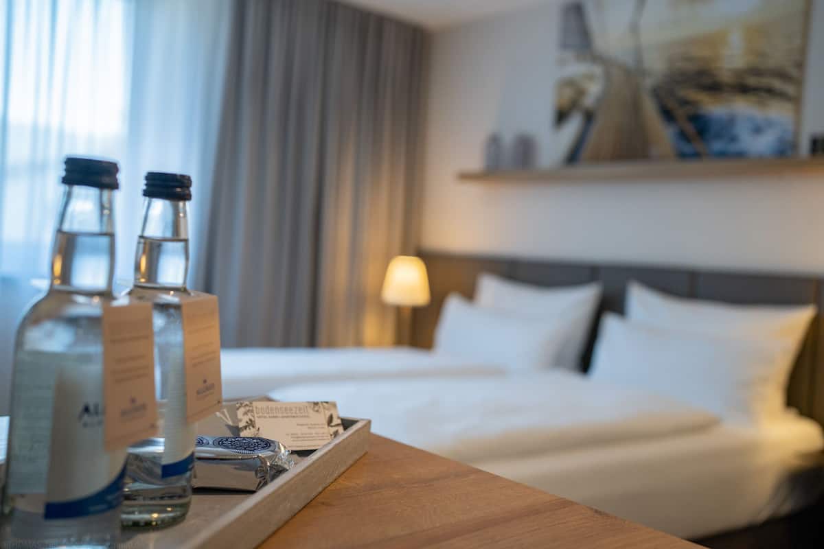 A hotel room with a bed, a bedside table with bottles and a tray, and a lamp.