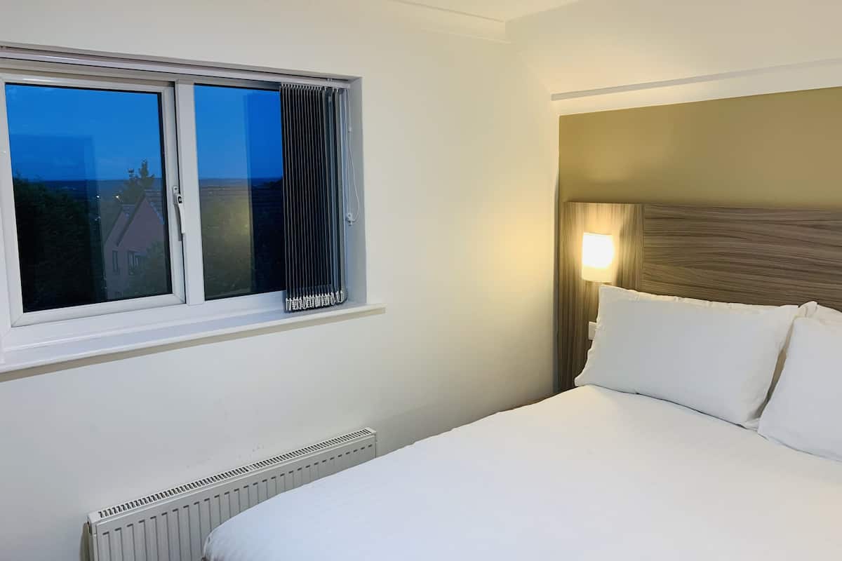 Habitación de hotel con cama, ventana con cortinas, radiador y lámpara fijada a la pared.