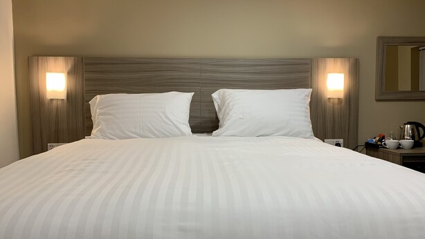 A neatly made bed with two pillows, a wooden headboard, and bedside lamps. - De Regency Style Hotel (Redditch)