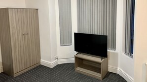 A room with a large wardrobe, a flat-screen TV on a wooden stand, and a window with vertical blinds. - De Regency Style Hotel (Redditch)