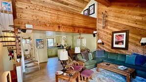 A cozy log cabin interior with a wooden staircase, a dining area, a green sofa, and a kitchen visible through an open doorway. - Bright + Beautiful, steps to Old Mammoth + Shuttle (Mammoth Lakes)