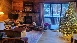 A cozy cabin interior with a stone fireplace, wooden furniture, a Christmas tree, and a view of a snowy landscape. - Bright + Beautiful, steps to Old Mammoth + Shuttle (Mammoth Lakes)