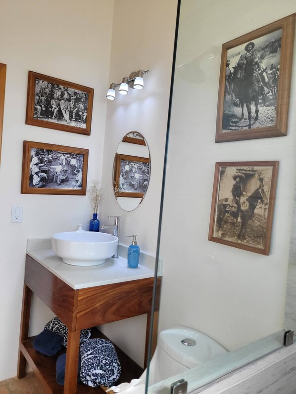 Un baño con lavamanos blanco, tocador de madera y espejo. Hay cuatro fotografías en blanco y negro en la pared.
