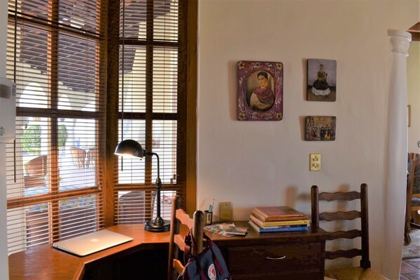 Una sala de estudio con un escritorio de madera, una computadora portátil, una lámpara y una silla. Hay libros y una mochila sobre el escritorio.