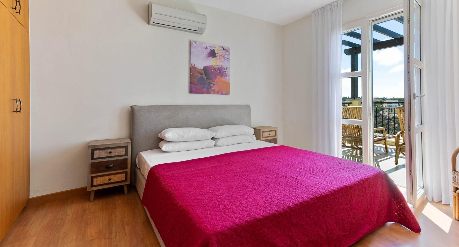 A bedroom with a bed, nightstands, an air conditioner, a painting, and a balcony with outdoor furniture.