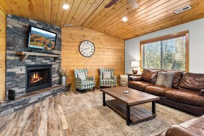 A cozy living room with a fireplace, a television mounted on a stone wall, a large clock, and a leather sofa.