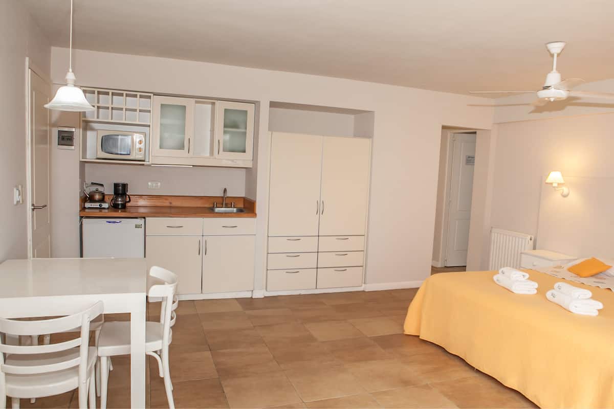 A compact hotel room with a kitchenette, a bed with yellow bedding, a ceiling fan, and a small table with chairs.