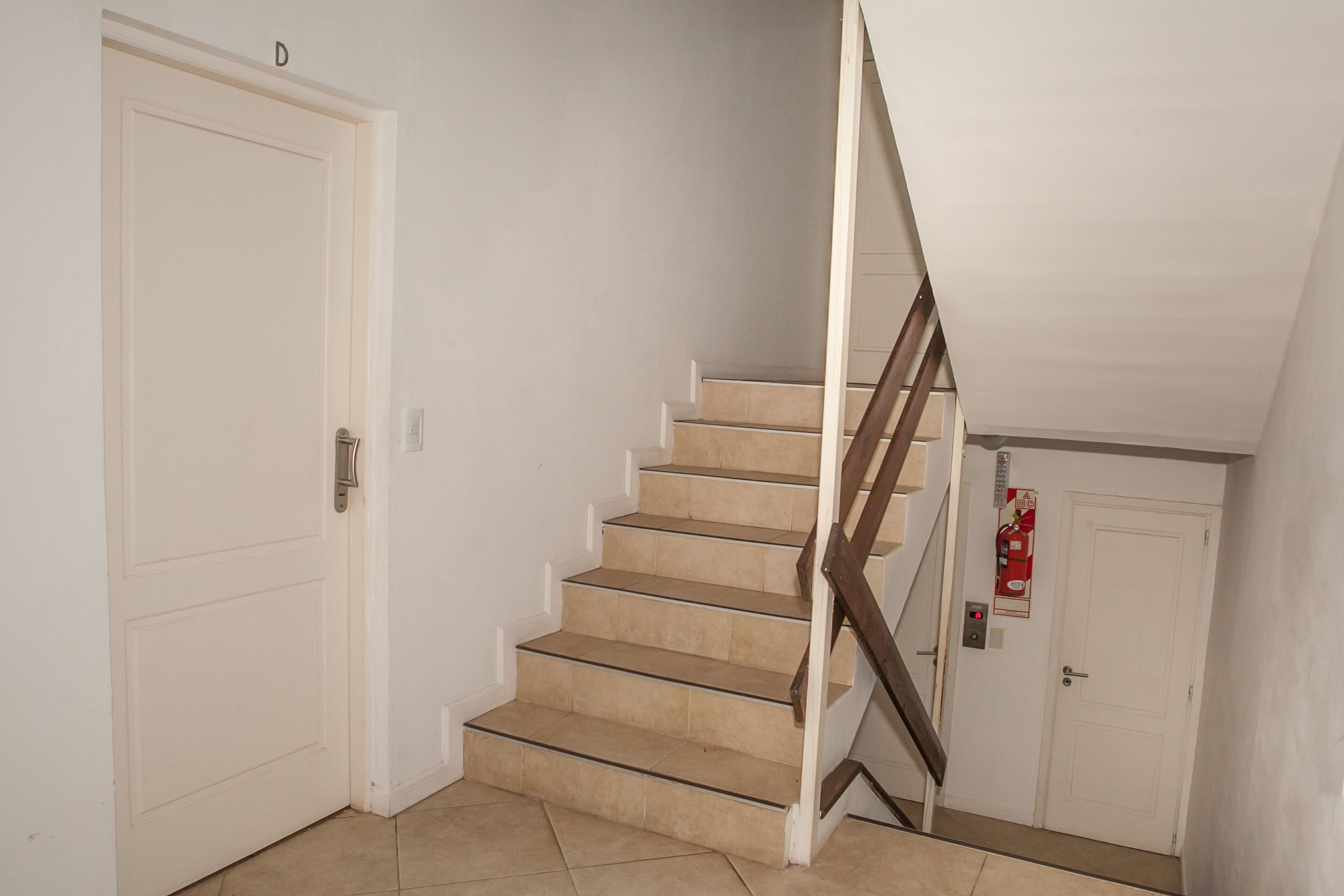 Een trap met beige treden en een houten leuning, die leidt naar een plateau met een witte deur waarop de letter 'D' staat.