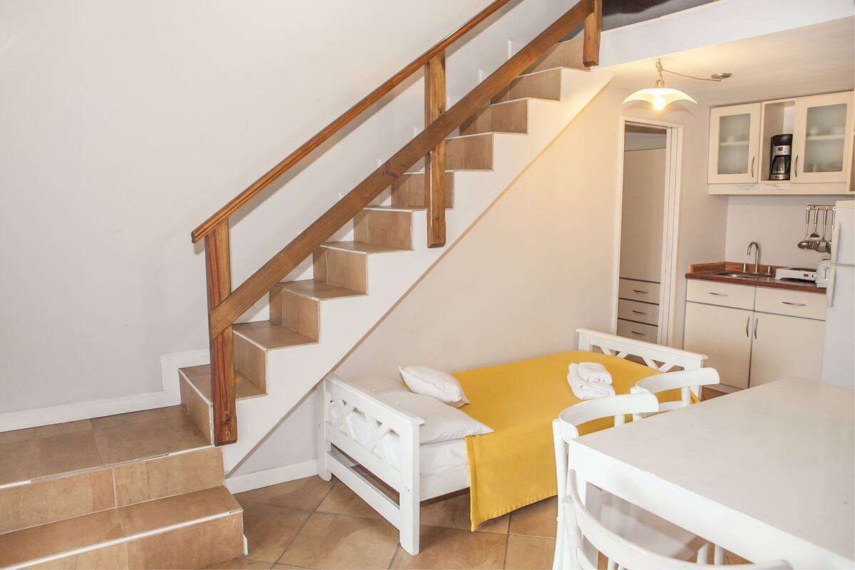 A bedroom with a bed, a wooden staircase, a kitchen area with cabinets, and a dining table.