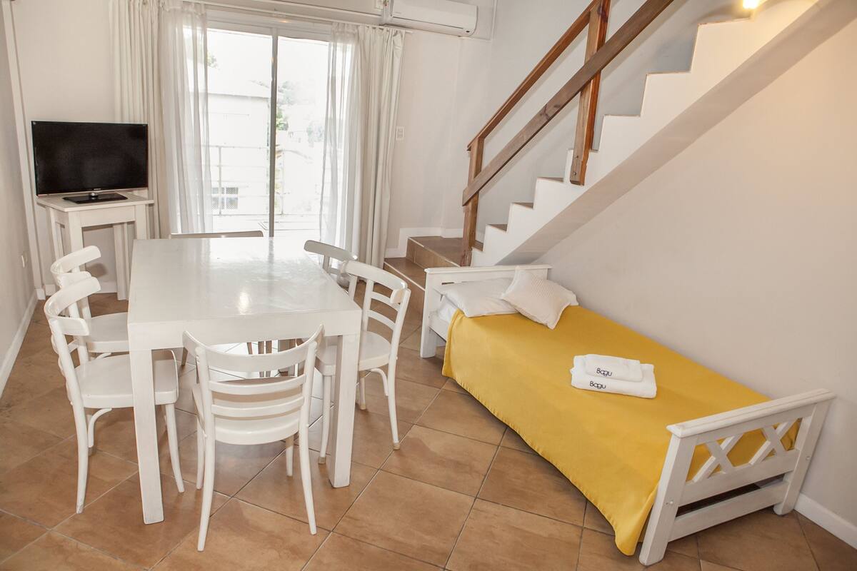 A room with a dining table, chairs, a sofa, a TV, and a staircase.