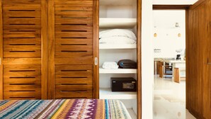 A bedroom with a wooden wardrobe, a bed with a patterned bedspread, and a white wall with a small shelf holding a radio and towels.