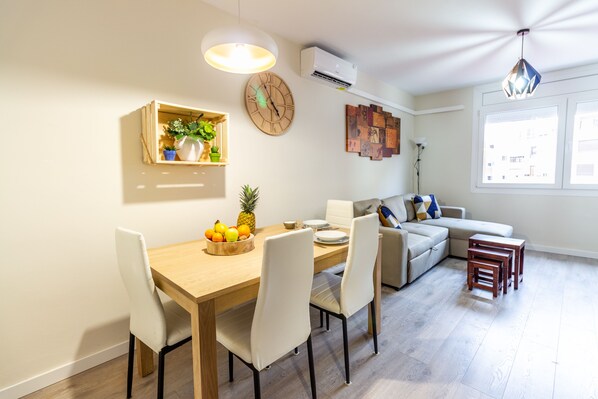 A dining area with a wooden table, white chairs, a fruit bowl, and a wall-mounted clock.