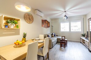 A modern living room with a dining area, featuring a wooden dining table, white chairs, a sofa, and a TV stand.