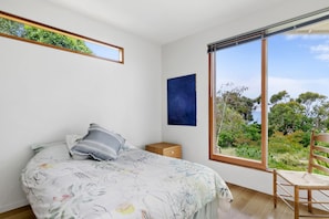 Ein Schlafzimmer mit einem Bett, einem Nachttisch, einem Sessel, einem großen Fenster und einem blauen Kunstwerk an der Wand.
