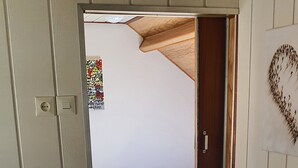 Un couloir étroit mène à une chambre à coucher qui contient un lit, une table de chevet avec une lampe et un mur orné d’un objet décoratif.
