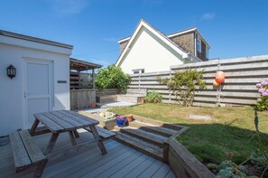 A wooden deck with picnic tables, a small garden with potted plants, and a white building with a wooden fence.