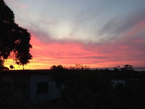 Un coucher de soleil avec un ciel aux teintes roses et violettes, un arbre et un bâtiment au premier plan.