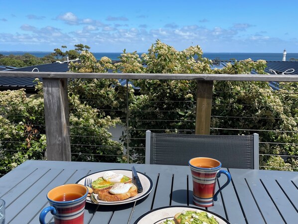 Une table dressée avec des aliments pour le petit-déjeuner et deux tasses, offrant une vue panoramique sur un paysage verdoyant avec des arbres et l’océan.
