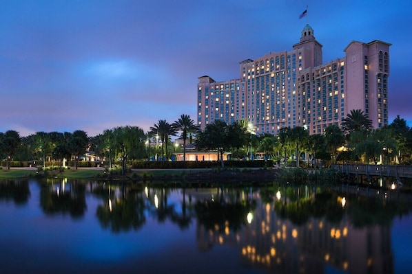 A large hotel with multiple floors and a central tower, surrounded by a body of water and palm trees.
