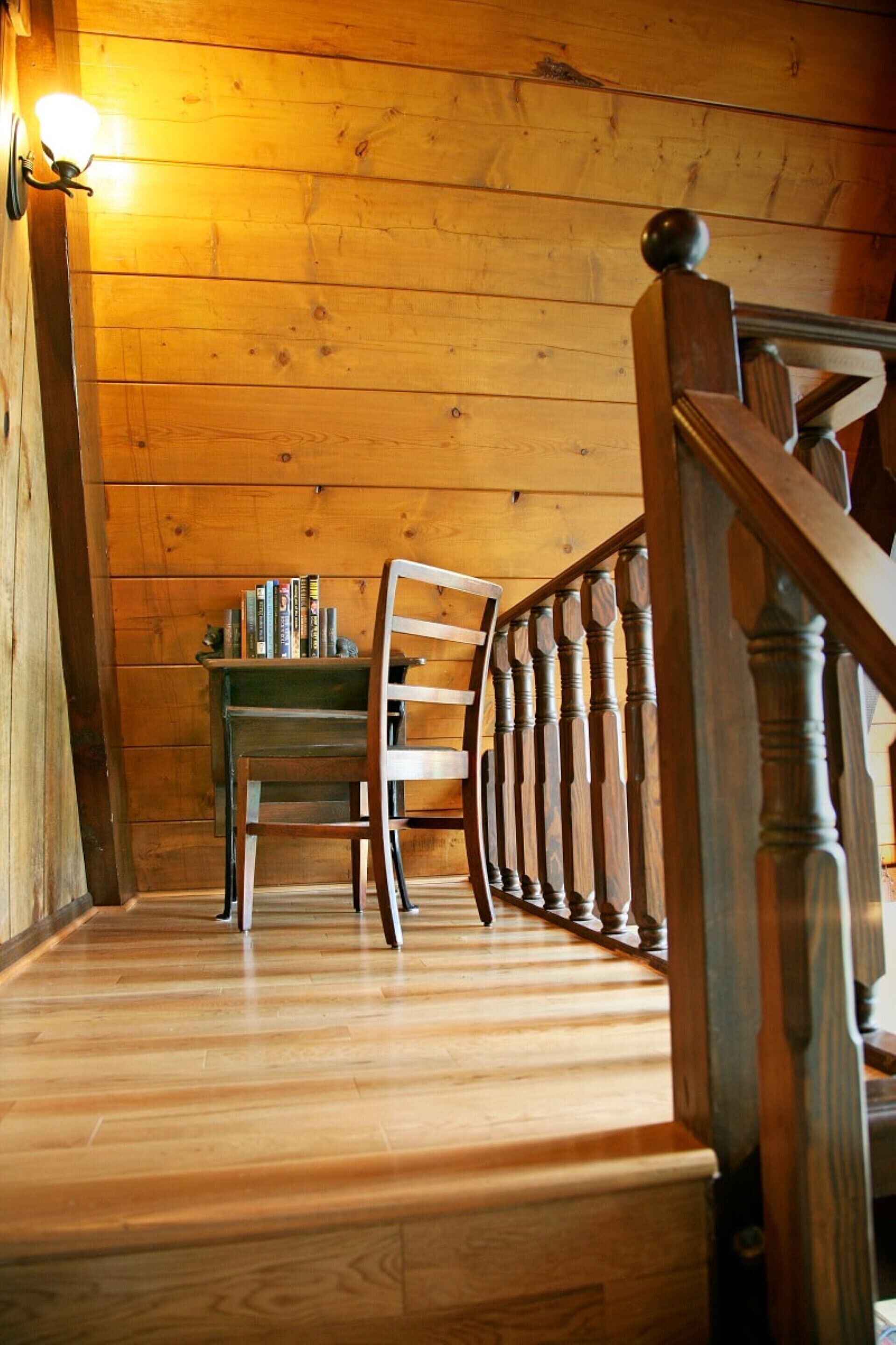 Una escalera de madera con una silla y libros, que lleva a una habitación con paredes de madera y una lámpara.