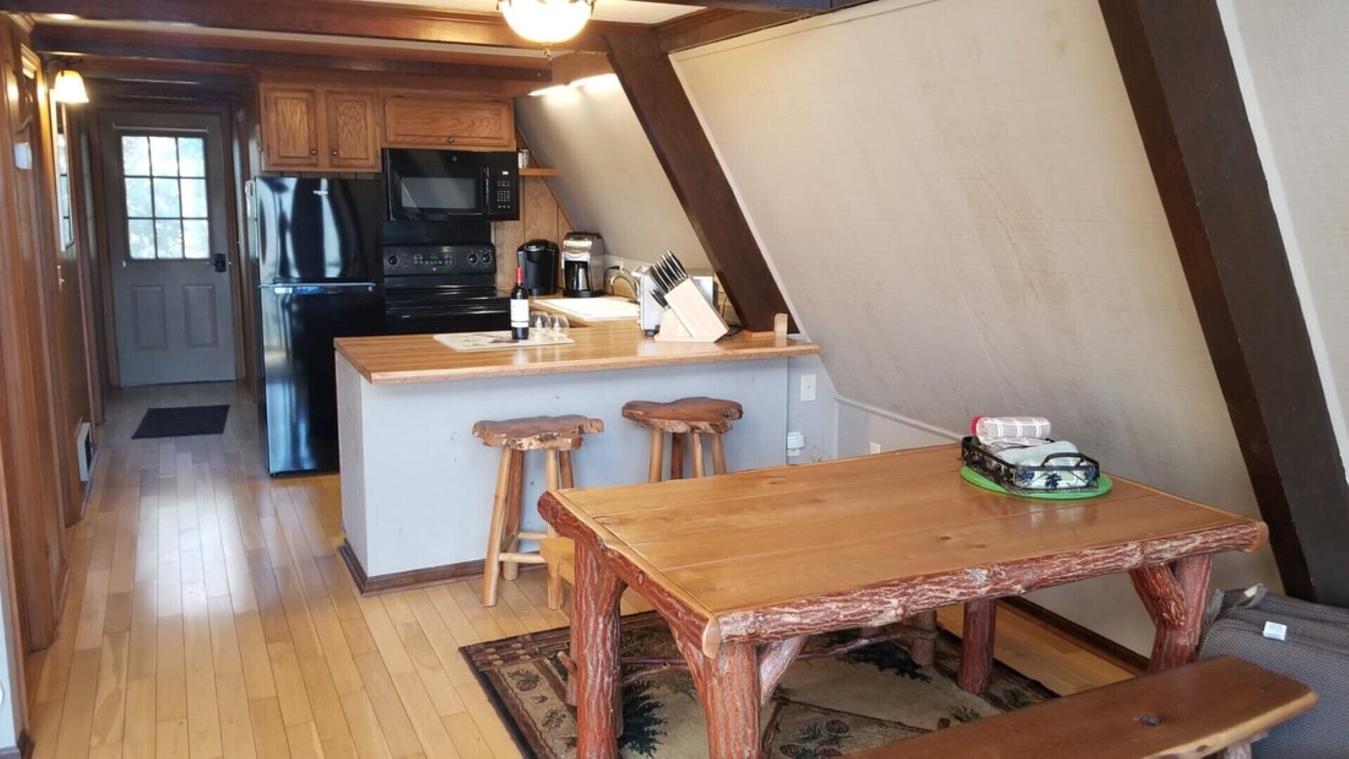 A kitchen with wooden cabinets, a microwave, and a coffee maker. There is a wooden table with two stools and a bench.