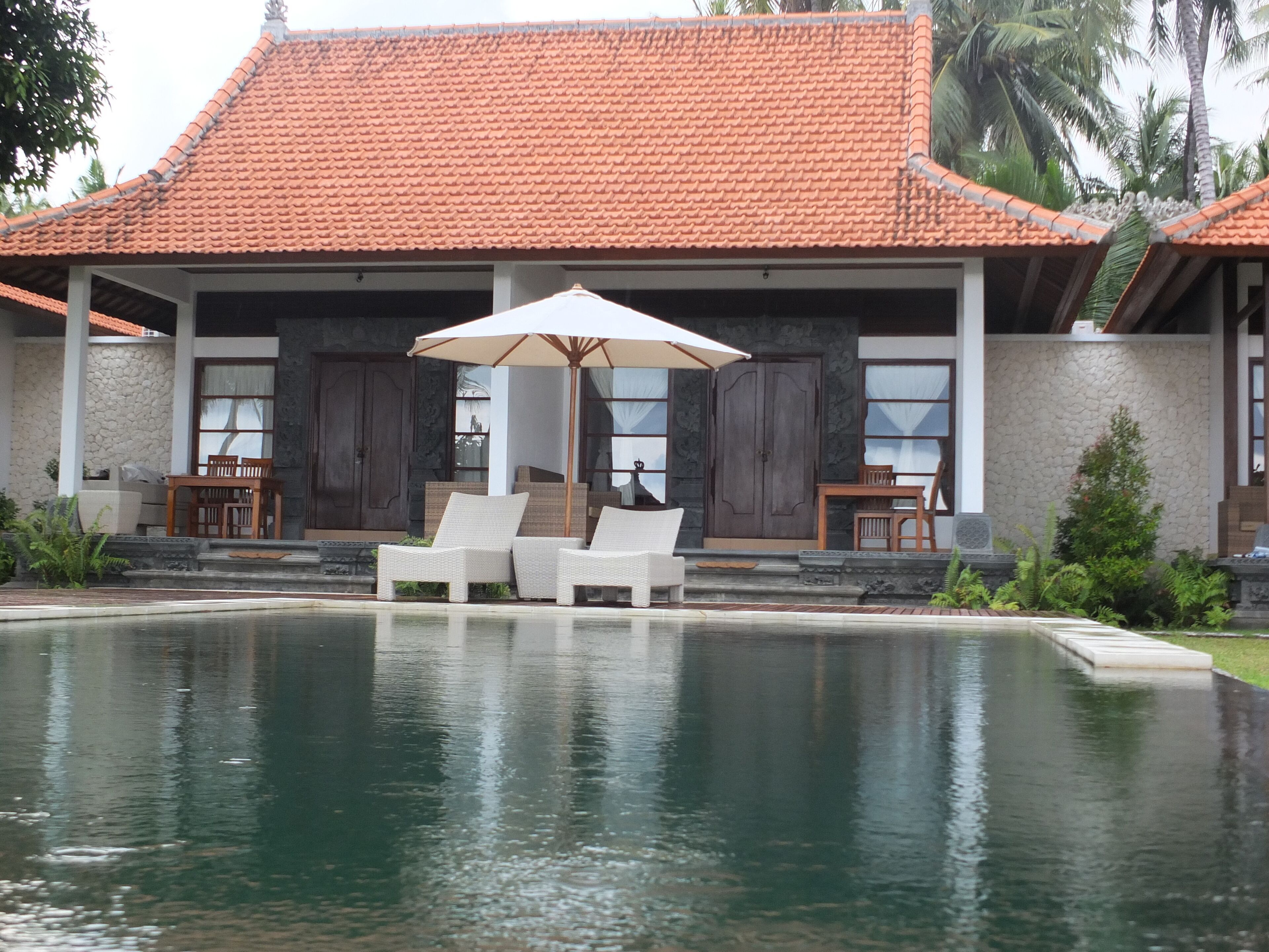 A poolside area with two white lounge chairs and a table under a large umbrella, surrounded by a building with a tiled roof and stone walls.