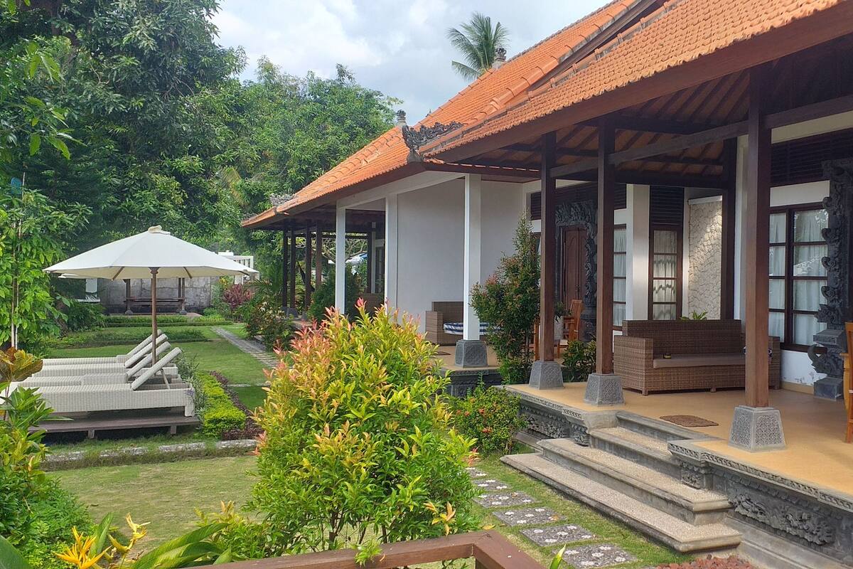 A traditional house with a tiled roof, a covered porch, and a garden with a lawn, a bench, and a sun umbrella.