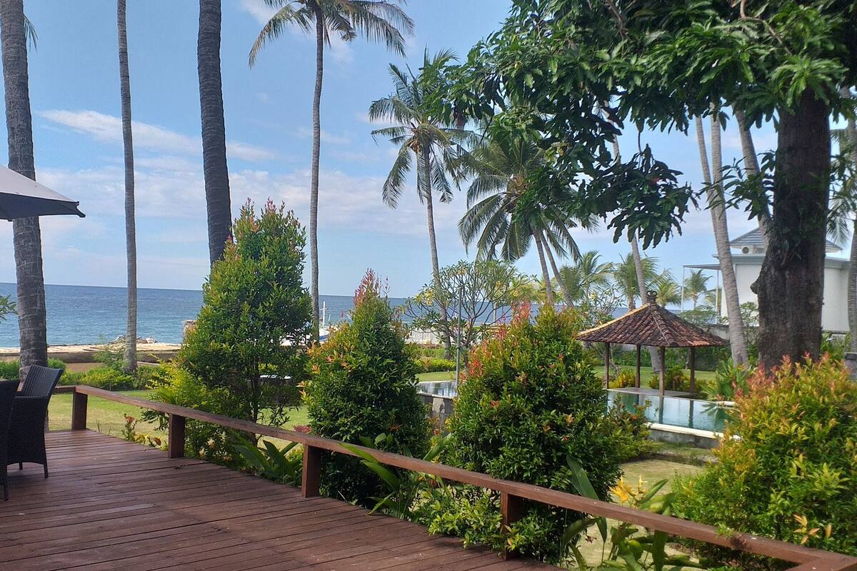 A wooden deck with a table and chairs, surrounded by lush greenery and palm trees, overlooking a pool and the ocean.