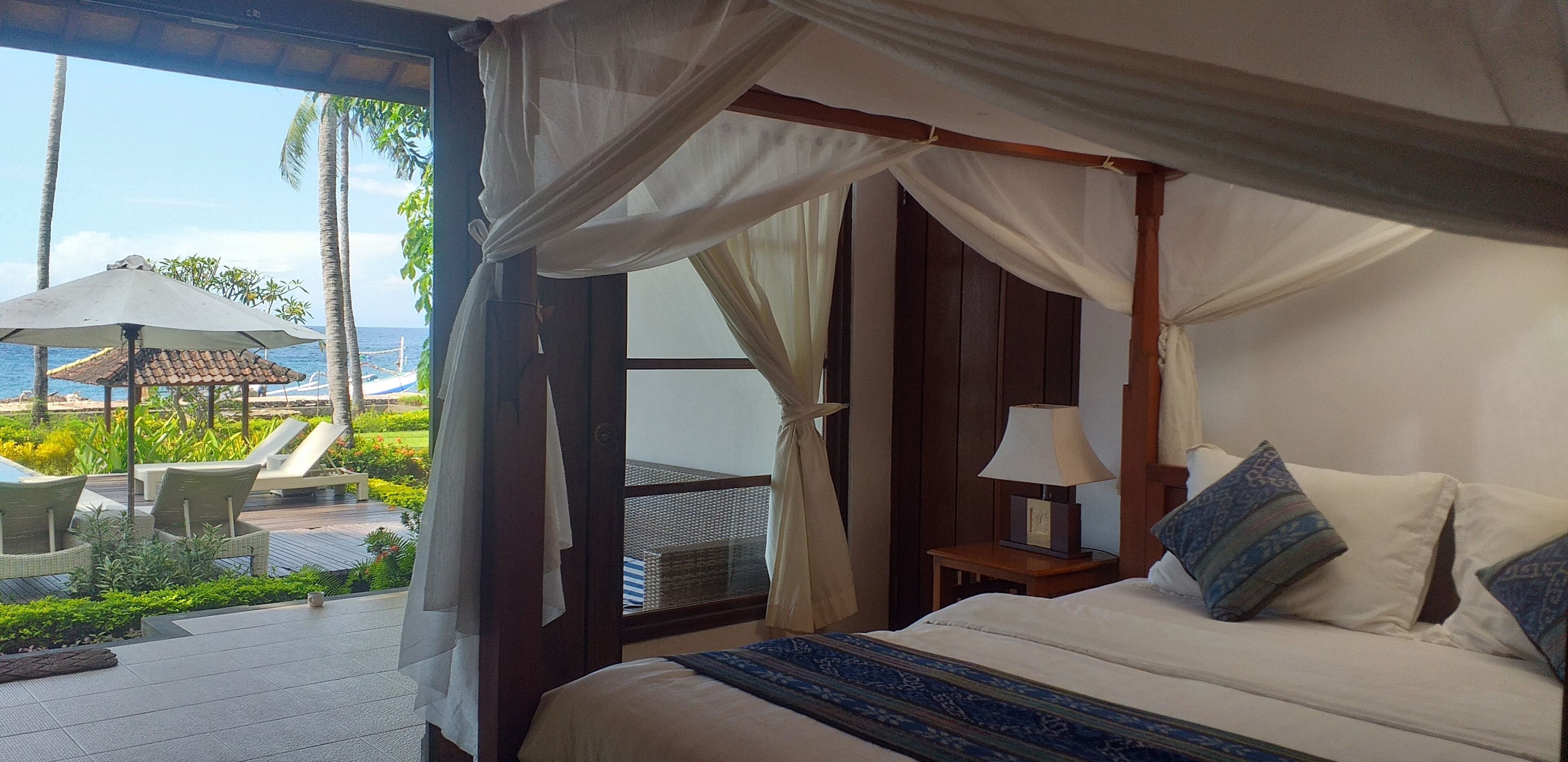 A bedroom with a bed, a nightstand, and a window overlooking a pool area with lounge chairs and umbrellas.