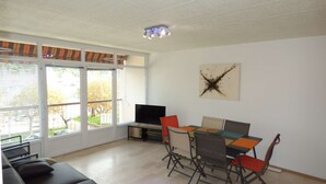 Ein Wohnzimmer mit Sofa, Fernseher, Esstisch und Stühlen. Ein großes Fenster mit Jalousien und eine Deckenleuchte sind ebenfalls vorhanden.
