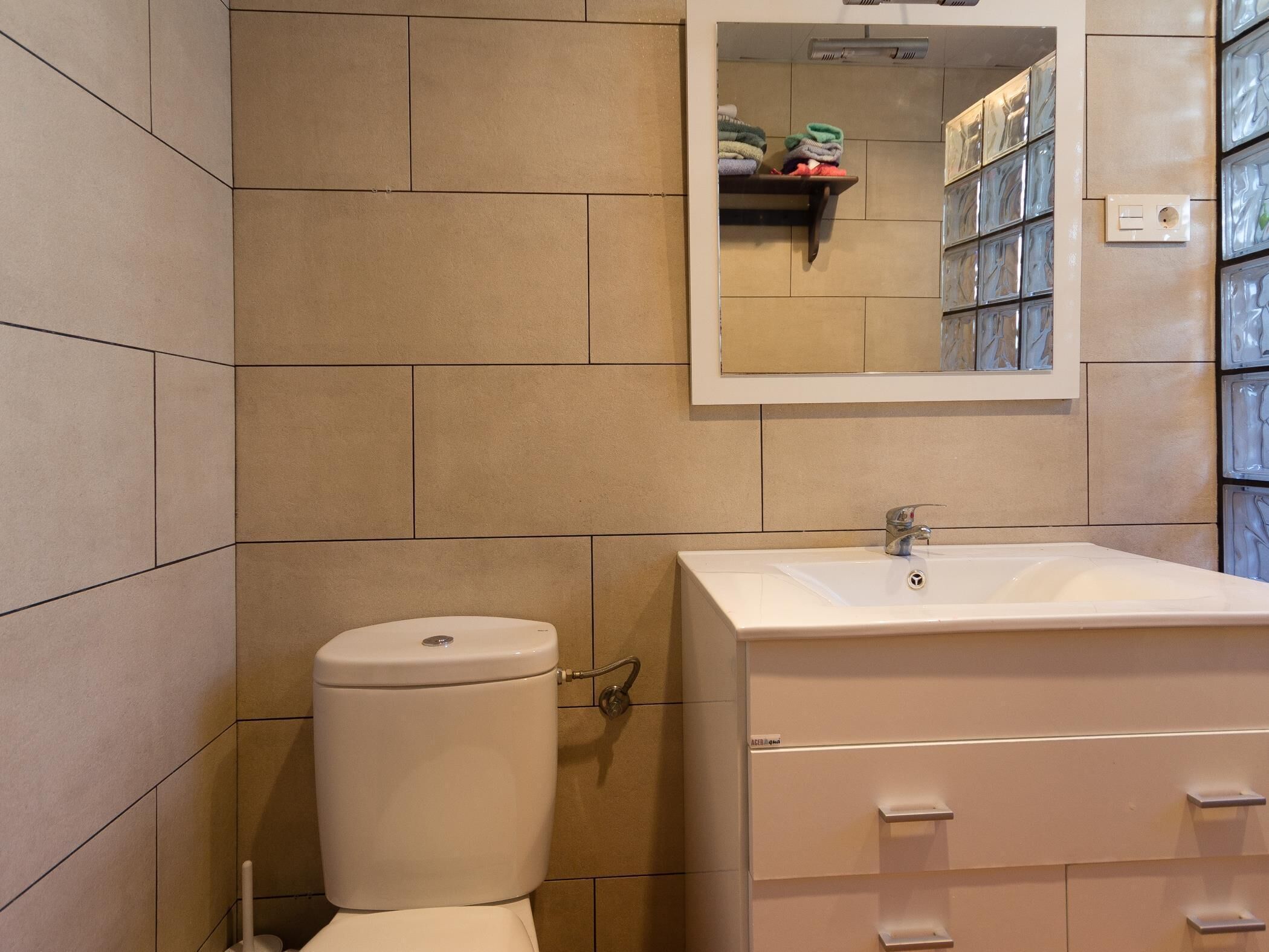 Un baño con inodoro, lavamanos y espejo. Hay un estante con toallas dobladas y un mueble pequeño con cajones.