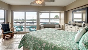 A bedroom with a large bed, a ceiling fan, a view of the water through large windows, and a balcony with chairs.