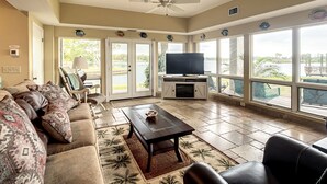 A spacious living room with a large TV, comfortable seating, and a view of a lake through large windows.