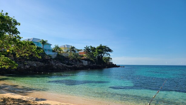 Une plage à l’eau turquoise et limpide, un rivage sablonneux et, en arrière-plan, des bâtiments bordés de palmiers.