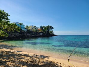 Ein Strand mit klarem türkisblauem Wasser, einem Sandstrand und Gebäuden mit Palmen im Hintergrund.
