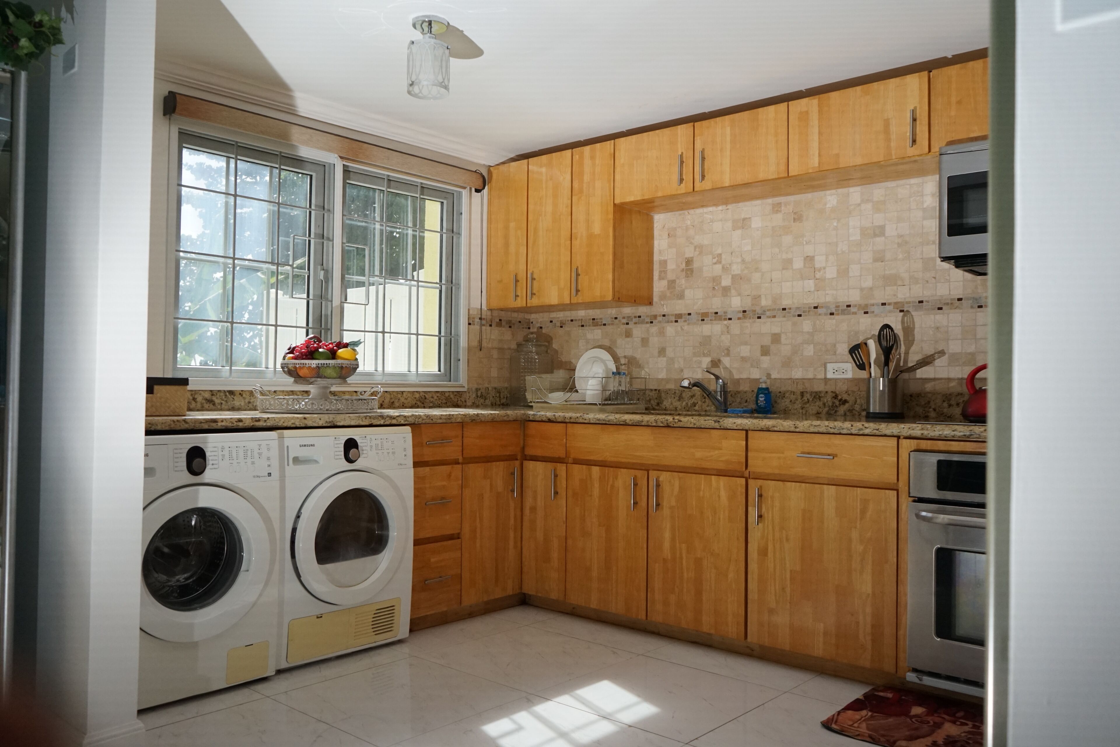 A kitchen with wooden cabinets, a built-in oven, a washing machine, and a window with a view of greenery.