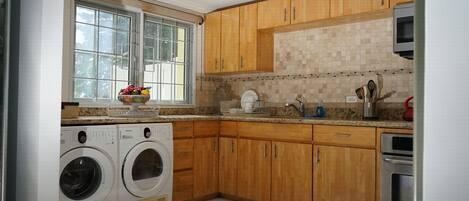 A kitchen with wooden cabinets, a built-in oven, a washing machine, and a window with a view of greenery.