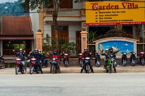 A group of motorcyclists posing in front of a building with a sign advertising Garden Villa, offering motorbike rentals, tour information, bus tickets, and accommodation. - Garden Villa - Hostel (Ha Giang 1)
