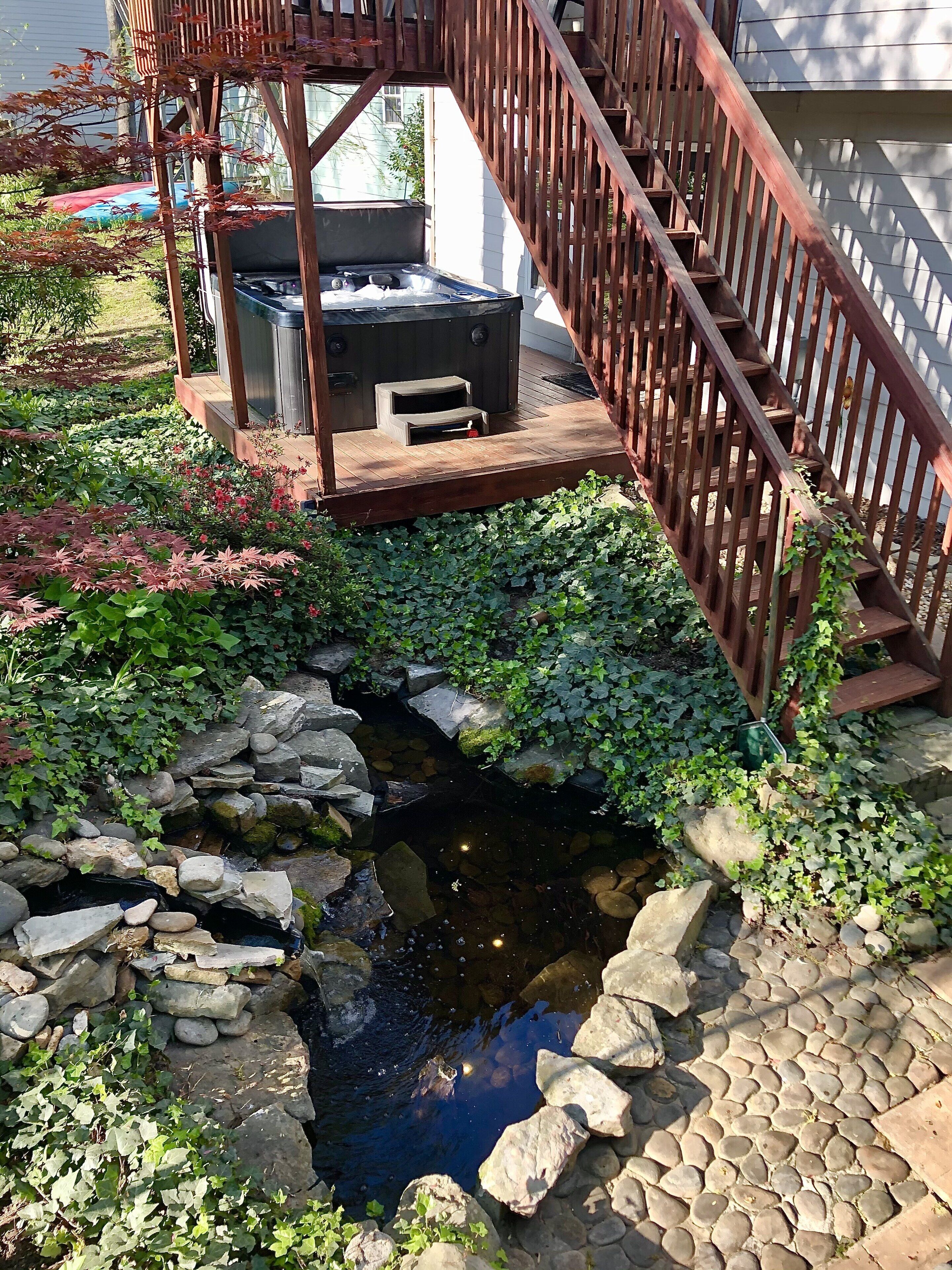 A wooden deck with a hot tub and a small pond surrounded by rocks and greenery.