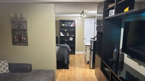 En kompakt bolig med sofa, tv, tekøkken og en gang der fører til en dør.