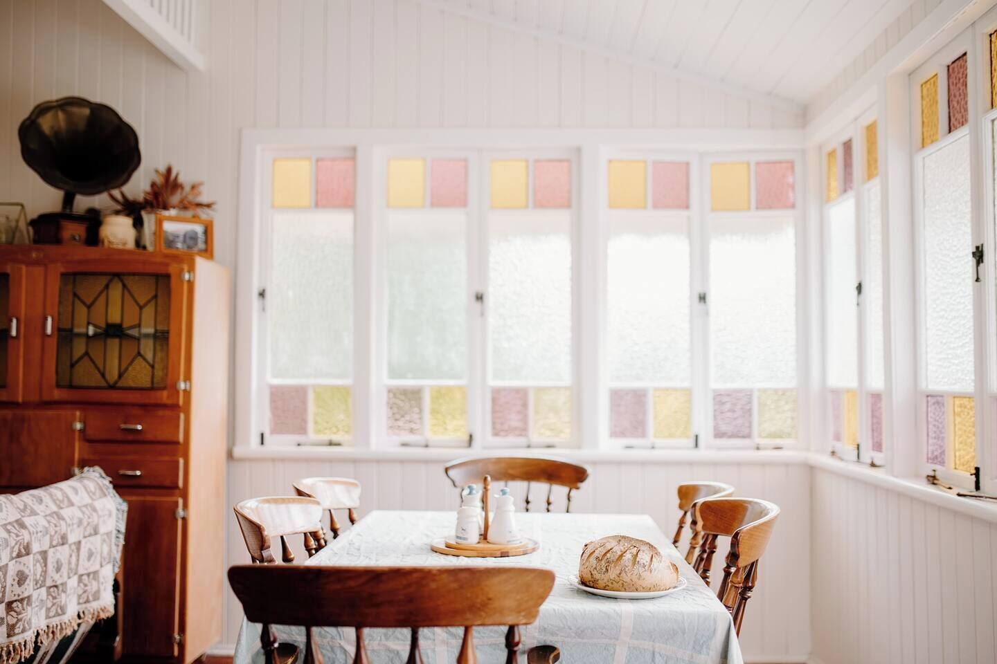 A dining room with a wooden table set for four, a cabinet with a decorative top, and a window with multicolored panes.