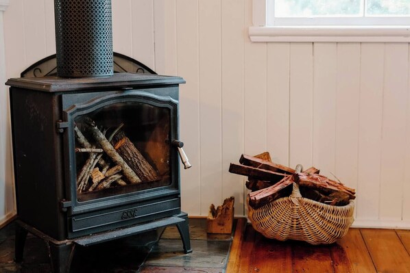 A wood-burning stove with logs inside, a wicker basket with firewood, and a window in the background.