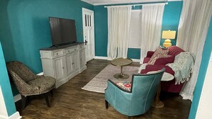 Uma sala de estar com paredes verde-água, piso de madeira, televisão sobre um móvel, sofá vermelho, poltrona com estampa de leopardo e uma mesa redonda pequena.