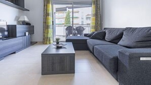 Ein modernes Wohnzimmer mit einem grauen Sofa, einem Couchtisch und einem Fernseher auf einem Holzkasten. Durch das Fenster ist ein Balkon mit gelben Vorhängen zu sehen.
