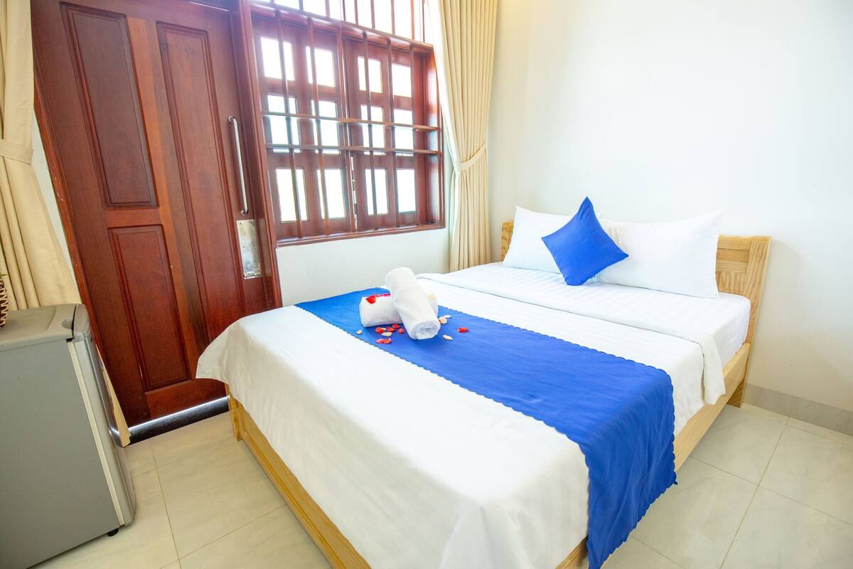 A hotel room with a bed, a wooden door, a window with bars, and a small refrigerator.