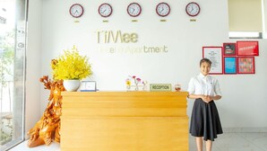 A hotel reception desk with a receptionist, a clock, and a sign that reads "TiMee Hotel & Apartment." - TiMee Hotel & Apartment (Nam Nha Trang)
